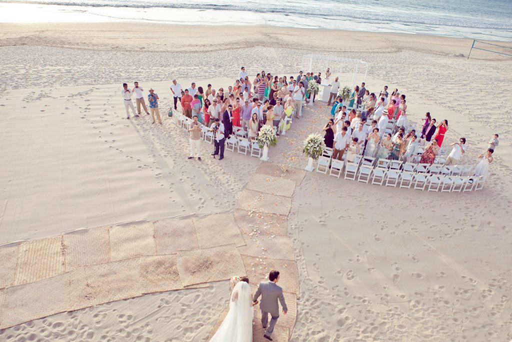 A bride and groom walk down a sandy aisle toward guests seated in a semicircle at their Beach Destination Wedding, with the ocean visible in the background at Hard Rock Hotel Puerto Vallarta, Mexico.