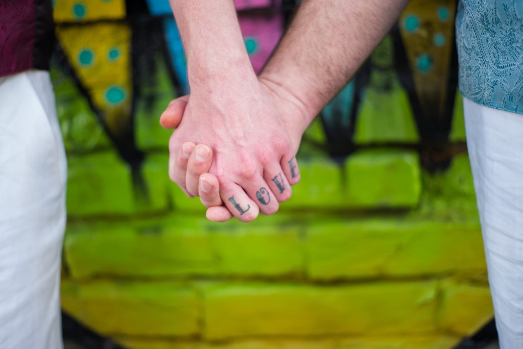 Two people hold hands in front of a colorful, graffiti-style wallâ€”one with LOVE tattooed across their fingers. Captured by a Puerto Vallarta gay wedding photographer, only their hands and part of their arms are visible.