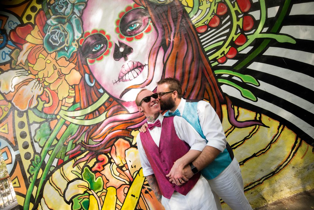 Two men in bright vests and bow ties embrace and smile before a vibrant Day of the Dead mural. Captured by a Puerto Vallarta gay wedding photographer, this joyful moment radiates love and color.
