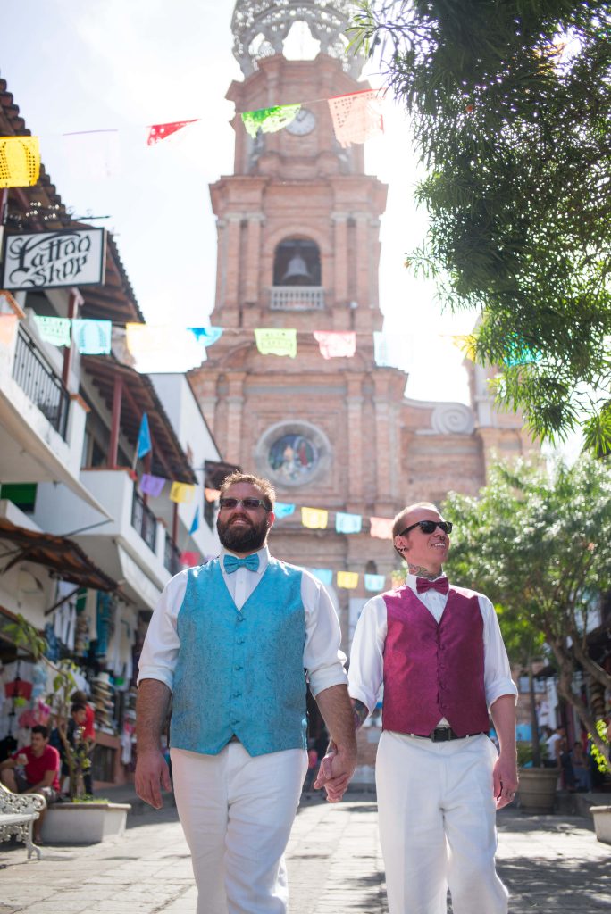 Two men dressed in formal vests and bow ties walk hand in hand down a sunlit street, with colorful banners overhead and a historic church behindâ€”an unforgettable moment perfectly captured by a Puerto Vallarta gay wedding photographer.