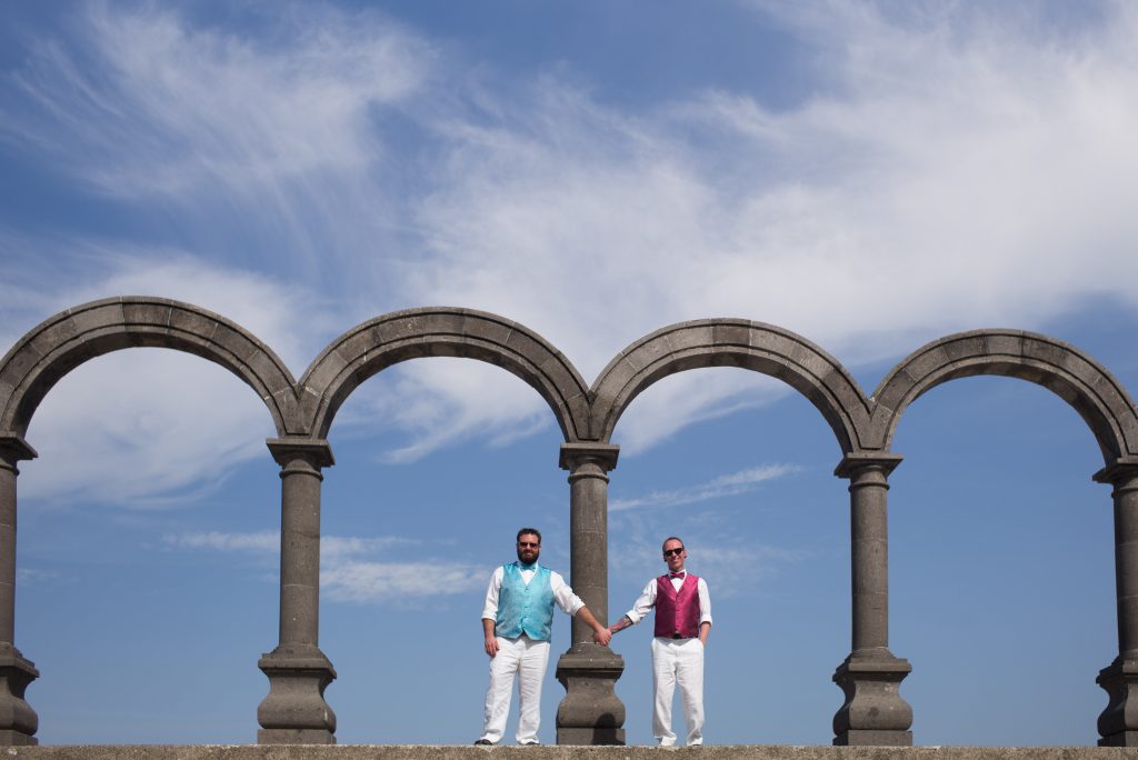 Two men stand holding hands under stone arches at Le Kliff against a bright blue sky, dressed in white pants and colorful vests. This joyful, celebratory scene is perfectly captured by a Puerto Vallarta Gay Wedding Photographer.