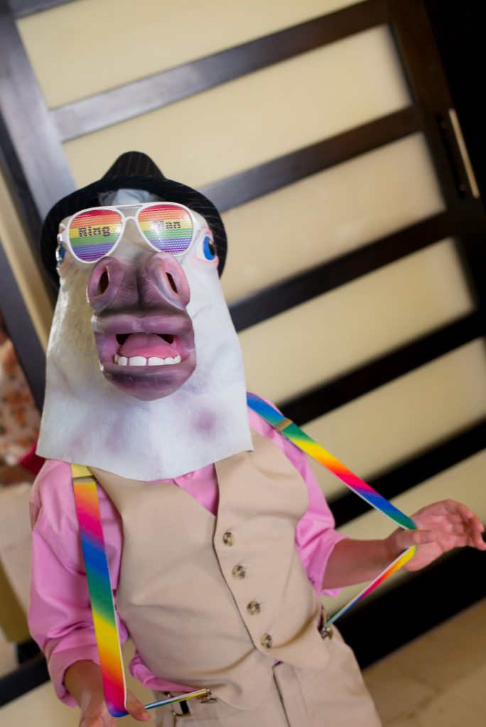 A person wearing a horse mask, rainbow sunglasses with â€œRing Manâ€ text, a tan vest over a pink shirt, and rainbow suspenders stands indoors holding out the suspendersâ€”ready to celebrate at a Puerto Vallarta gay wedding photographerâ€™s event.