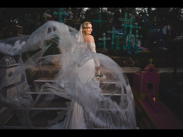 A bride in a white dress and veil smiles over her shoulder while standing outdoors at an Xcaret Park Mexico wedding, holding a bouquet. Colorful crosses and structures are visible as her long veil flows dramatically in the wind.