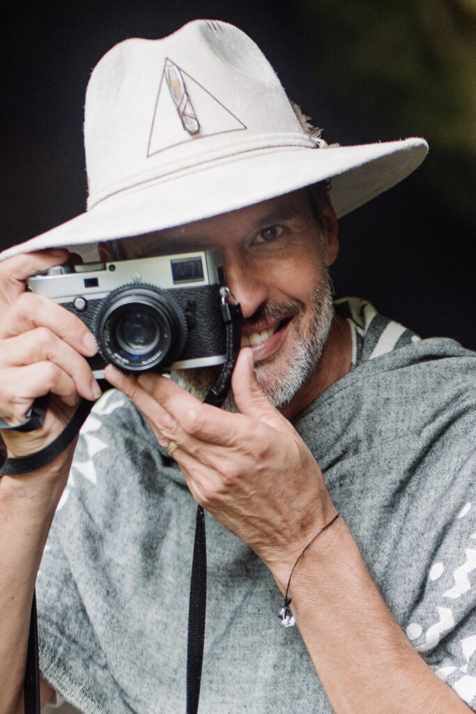A smiling man in a wide-brimmed hat and poncho holds a vintage camera up to his eye, capturing moments like the best photographer in Riviera Maya at Hotel Xcaret Weddings.