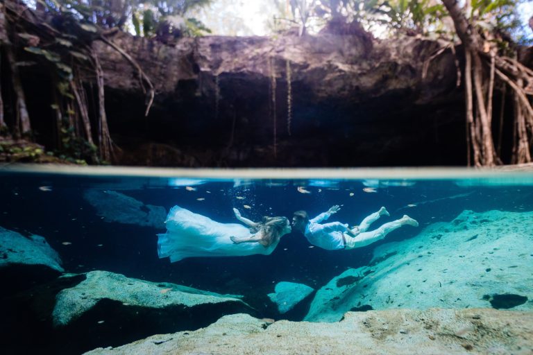 A bride and groom in wedding attire swim underwater in a clear cenote near Cancun, Mexico, surrounded by rocks and tree roots, with sunlight filtering from aboveâ€”captured by a Playa del Carmen Wedding Photographer.