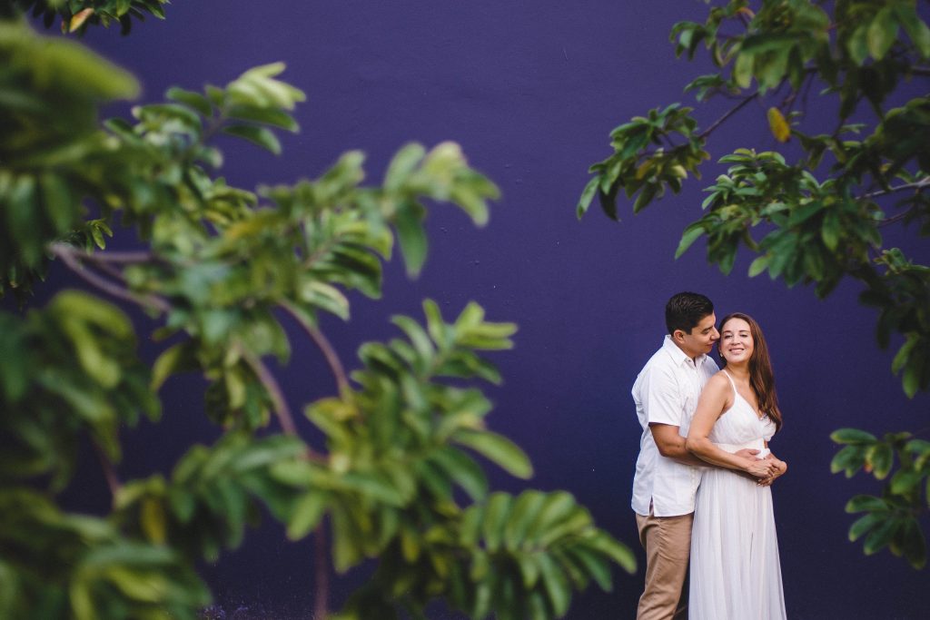 A couple stands embracing and smiling before a deep purple wall at Hotel Xcaret Arte, framed by green leafy branches. The woman wears a white dress, while the man stands behind her in a white shirt and khaki pantsâ€”capturing Riviera Maya romance.