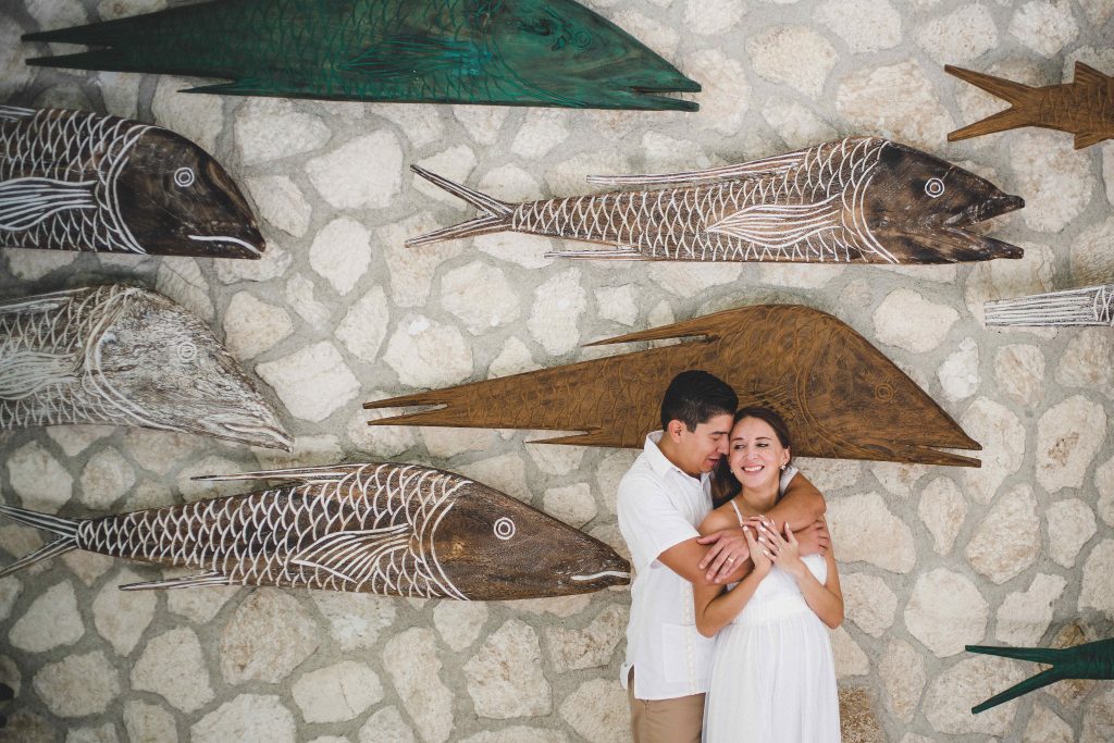 A couple embraces and smiles in front of a stone wall decorated with large, carved wooden fish art at Hotel Xcaret Arte in Riviera Maya. The man hugs the woman from behind as she holds his arms, both dressed in white.
