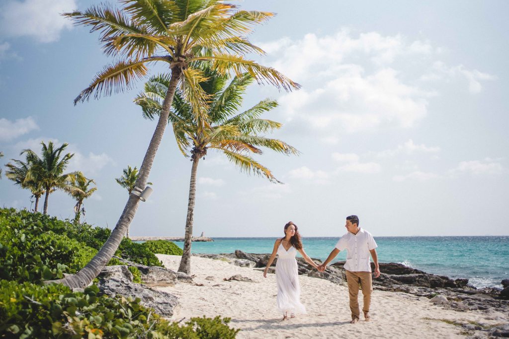 A couple walks hand in hand along a sandy Riviera Maya beach lined with palm trees, with clear blue ocean water and a bright sky in the background.