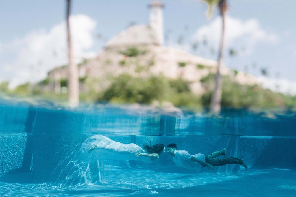 Two people swim underwater in a pool at Hotel Xcaret Arte, holding hands. The waterline splits the image, showing blurred scenery with palm trees and a stone building above the surface. The Riviera Maya scene is bright and serene.