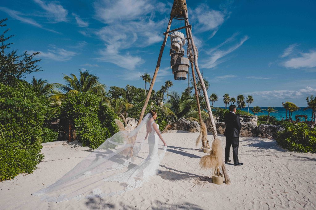 A bride in a white dress and veil walks toward a groom on a sandy Riviera Maya beach, surrounded by palm trees and greenery, with blue sky and ocean in the background. A decorative wooden structure stands at the center of this Hotel Xcaret Arte scene.