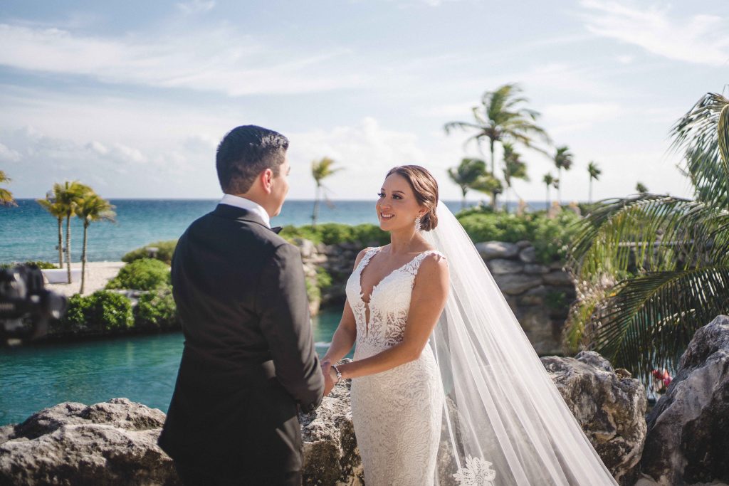 A bride and groom stand outdoors at Hotel Xcaret Arte, holding hands by the water. With palm trees, blue sky, and the ocean framing their Catholic wedding, she wears a white dress and veil; he is dressed in a black suit in Riviera Maya.