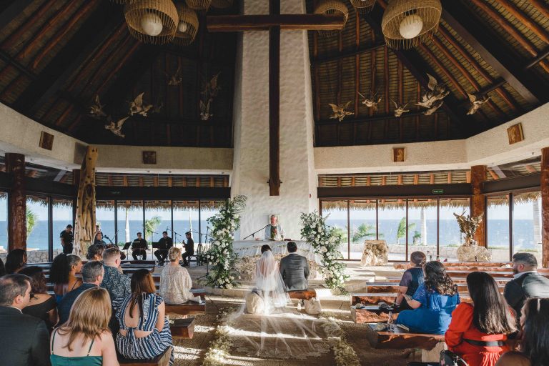 A bride and groom sit at the altar of a sunlit, open-air chapel with wooden beams and high ceilings, surrounded by guests on benches and tropical floral arrangements in the heart of Riviera Maya at Hotel Xcaret Arte.