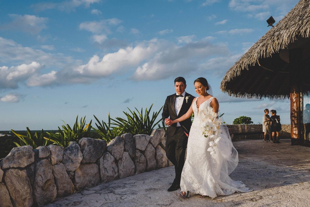 A bride in a white dress holding a bouquet walks arm-in-arm with her groom along a stone path at Hotel Xcaret Arte, surrounded by lush greenery, a thatched roof building, and blue skiesâ€”a stunning Riviera Maya Catholic wedding scene.