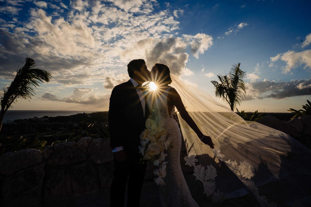 A bride and groom share a kiss at sunset during their Catholic wedding at Hotel Xcaret Arte, silhouetted against the Riviera Maya sky. The brideâ€™s veil blows in the wind as sunlight creates a starburst amid palm trees and clouds.