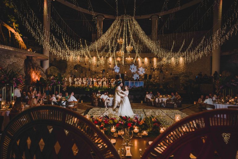 A bride and groom share their first dance under a canopy of string lights at an outdoor wedding reception, surrounded by guests seated at decorated tables with flowers and candles.