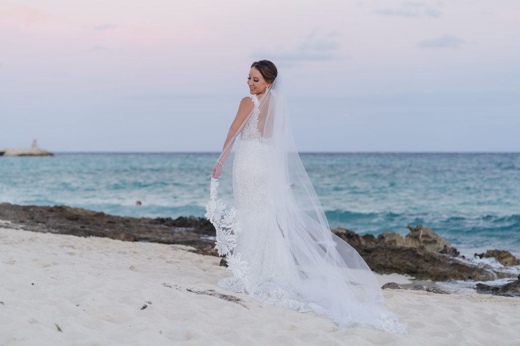 A bride in a white lace gown with a long veil stands on a sandy Riviera Maya beach, looking over her shoulder at the ocean. Rocky shoreline and gentle waves are in the background under a pastel sky.