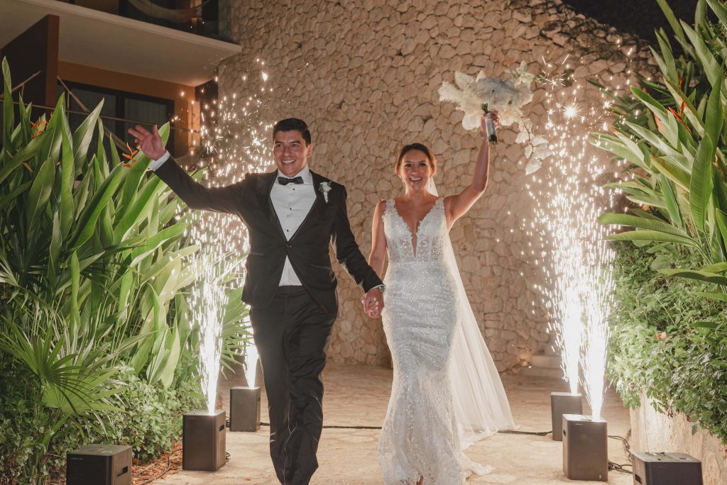 A bride and groom, holding hands and smiling, walk down an outdoor aisle at Hotel Xcaret Arte in Riviera Maya, surrounded by greenery and sparkling fireworks. The bride lifts a bouquet in celebration during their Catholic Wedding.