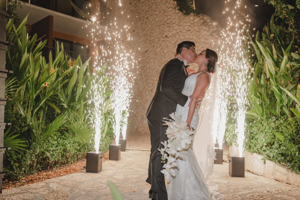 A bride and groom share a kiss outdoors at night at Hotel Xcaret Arte, surrounded by lush greenery, with sparkling fireworks erupting behind them, creating a celebratory and romantic Riviera Maya atmosphere.
