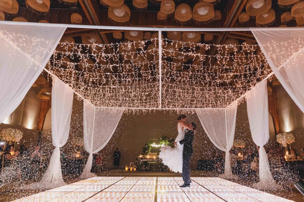A couple dances on a glowing floor under string lights at Hotel Xcaret Arte, surrounded by sheer white drapes and flowers, creating a magical and romantic Riviera Maya wedding atmosphere.