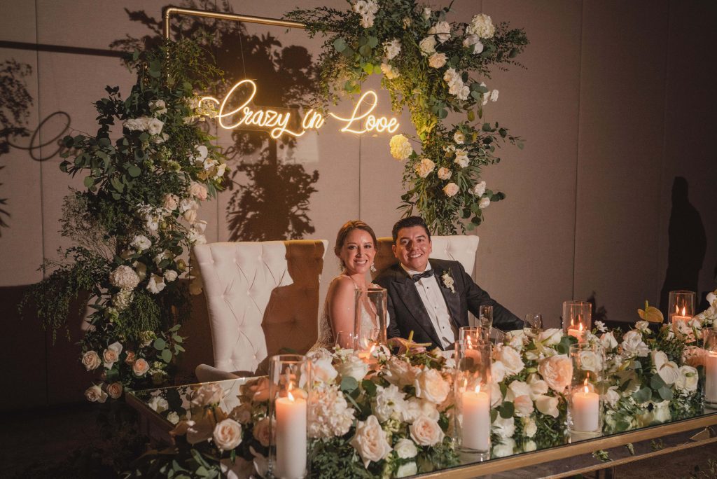 A bride and groom sit smiling at a flower-adorned table with lit candles. Behind them, a gold frame and greenery display a neon sign that reads â€œCrazy in Love.â€ The romantic, celebratory scene unfolds at Hotel Xcaret Arte in Riviera Maya.