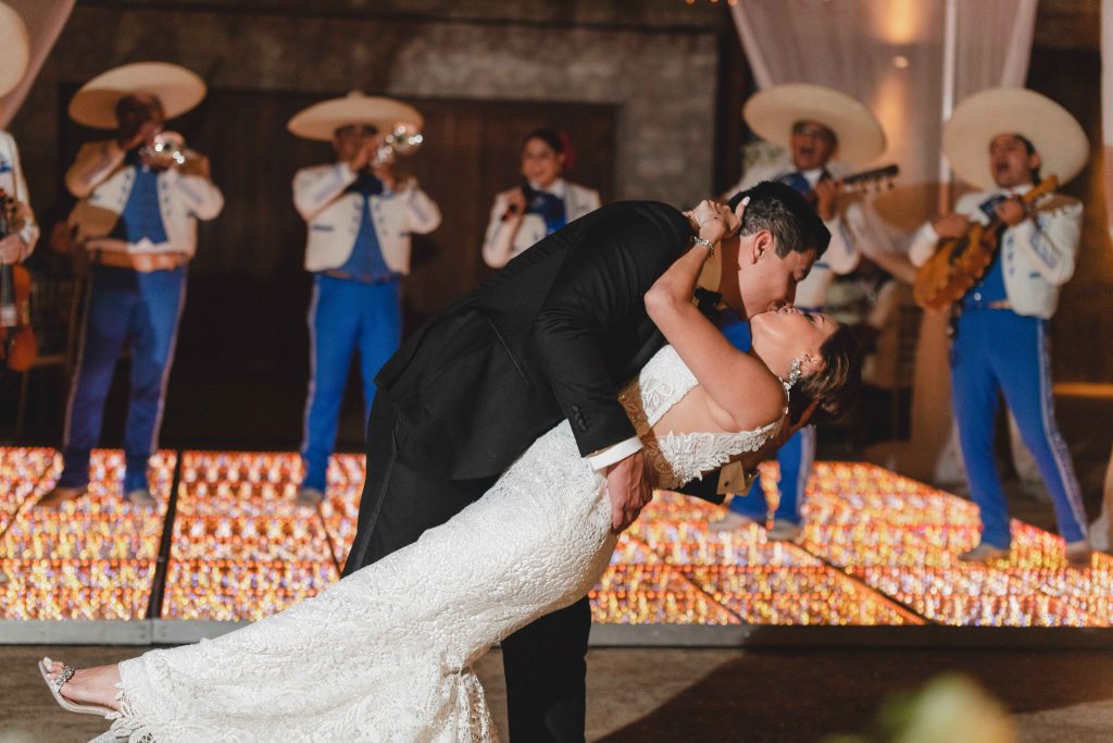 A bride and groom share a dramatic dip and kiss on a lit dance floor at Hotel Xcaret Arte, with a mariachi band dressed in blue and white performing in the background.