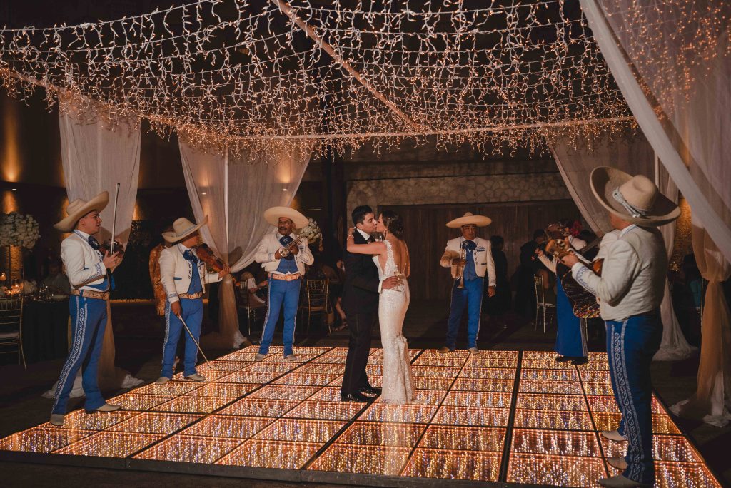 A bride and groom share a first dance on a lit-up floor under string lights at their Riviera Maya wedding, surrounded by a mariachi band in traditional outfits performing at their Hotel Xcaret Arte reception.