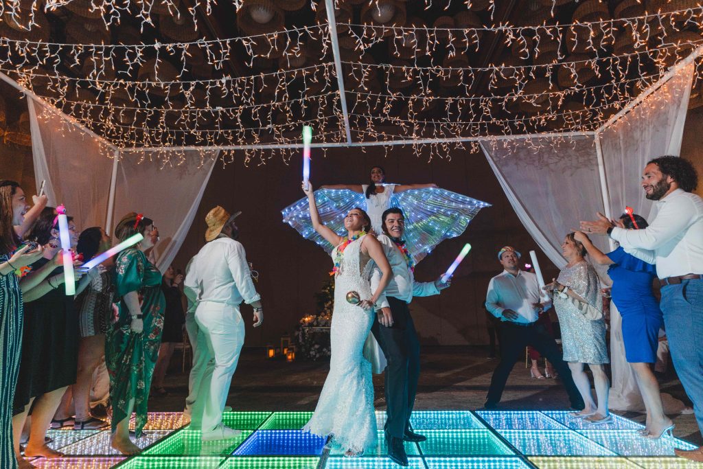 A bride and groom pose joyfully on a colorful, illuminated dance floor at Hotel Xcaret Arte in Riviera Maya, surrounded by cheering guests holding glow sticks, with festive string lights and a performer with glowing wings behind them.