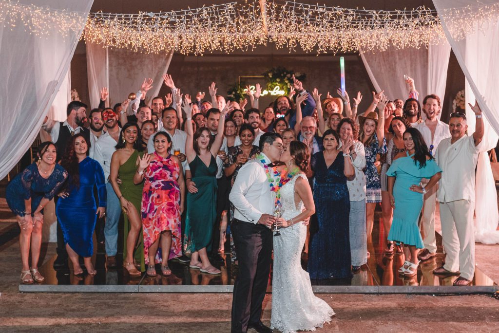 A bride and groom kiss at the front of a large group of guests, all smiling and celebrating at a festive indoor Catholic wedding with twinkling lights and draped white fabric in Riviera Mayaâ€™s stunning Hotel Xcaret Arte.