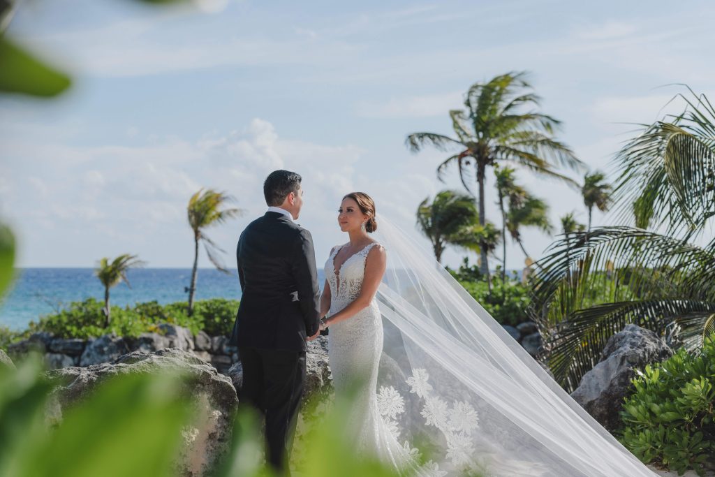 A bride and groom stand facing each other outdoors at Hotel Xcaret Arte, holding hands. The bride wears a long white dress and veil, and the groom a black suit. Palm trees and the Riviera Maya ocean are in the background under a clear blue sky.