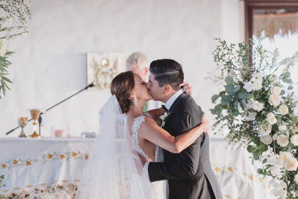 A bride and groom share a kiss at their Catholic wedding ceremony in Riviera Maya, standing before a decorated altar with white flowers and greenery. The bride wears a white dress and veil; the groom is in a black suit.