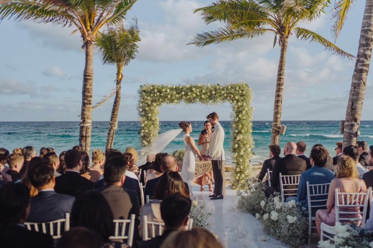 A bride and groom stand facing each other during an outdoor wedding ceremony by the ocean, under a floral arch and palm trees, with guests seated on white chairs watching.