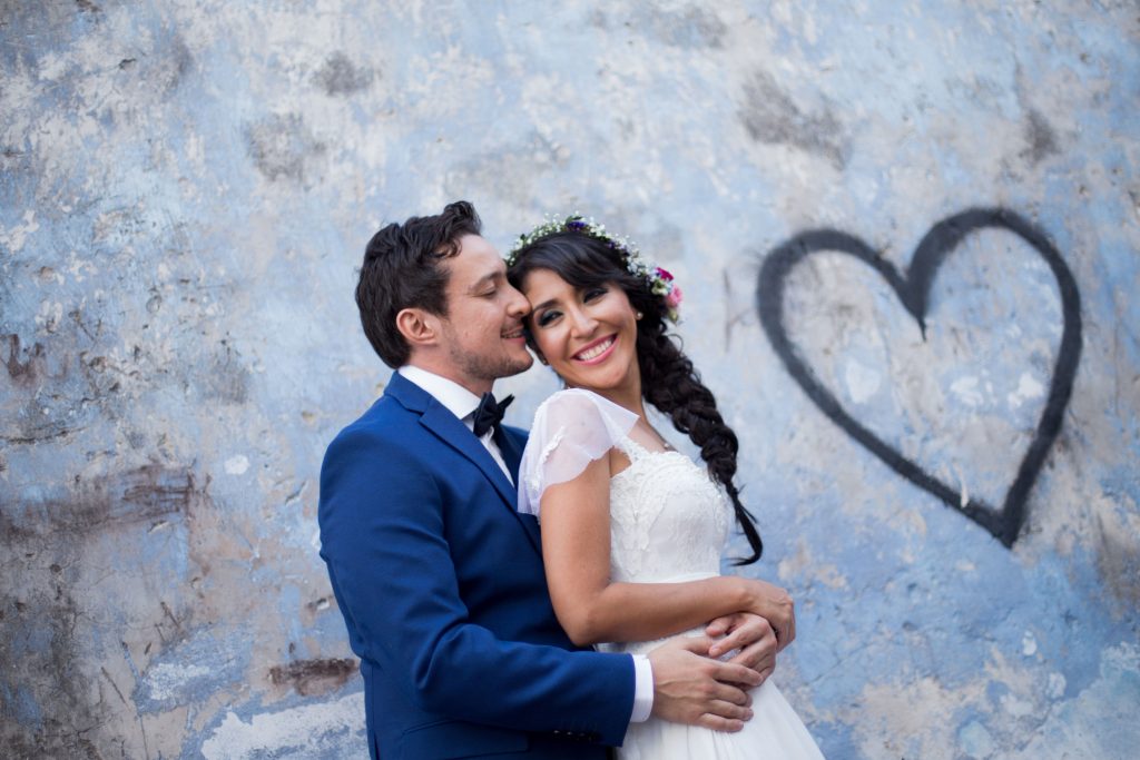 A groom in a blue suit kisses his smiling bride in a white dress and floral crown, standing before a weathered blue wall with black heart graffitiâ€”captured by your Yucatan Hacienda Wedding Photographer at Dzibikak.