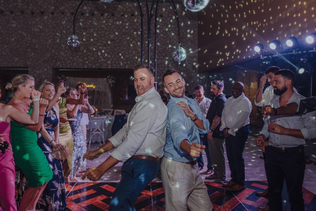 A group of people dance and laugh together at a lively party under disco lights, with two men in the center back-to-back, smiling as others enjoy the festive atmosphere around them.