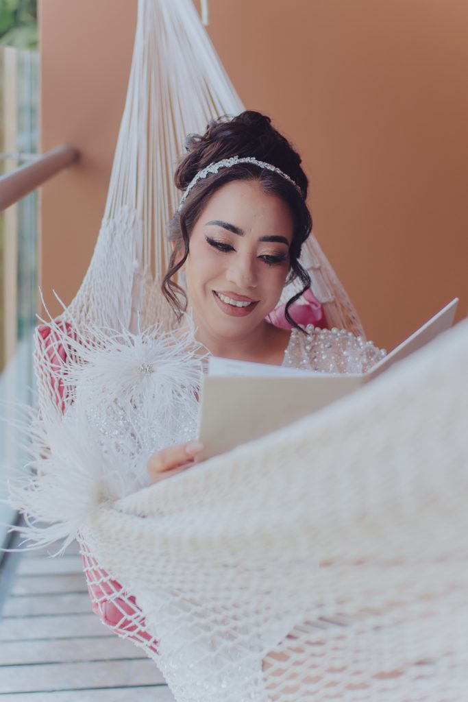 A smiling woman in a white dress sits in a white hammock, reading a card or letter. She has styled hair with a jeweled headband and is positioned against an orange wall.