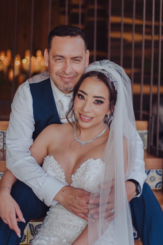 A smiling bride in a strapless white wedding gown and veil sits on her grooms lap. The groom, wearing a dark vest and white shirt, hugs her from behind as they pose on tiled steps. Candles glow softly in the background.