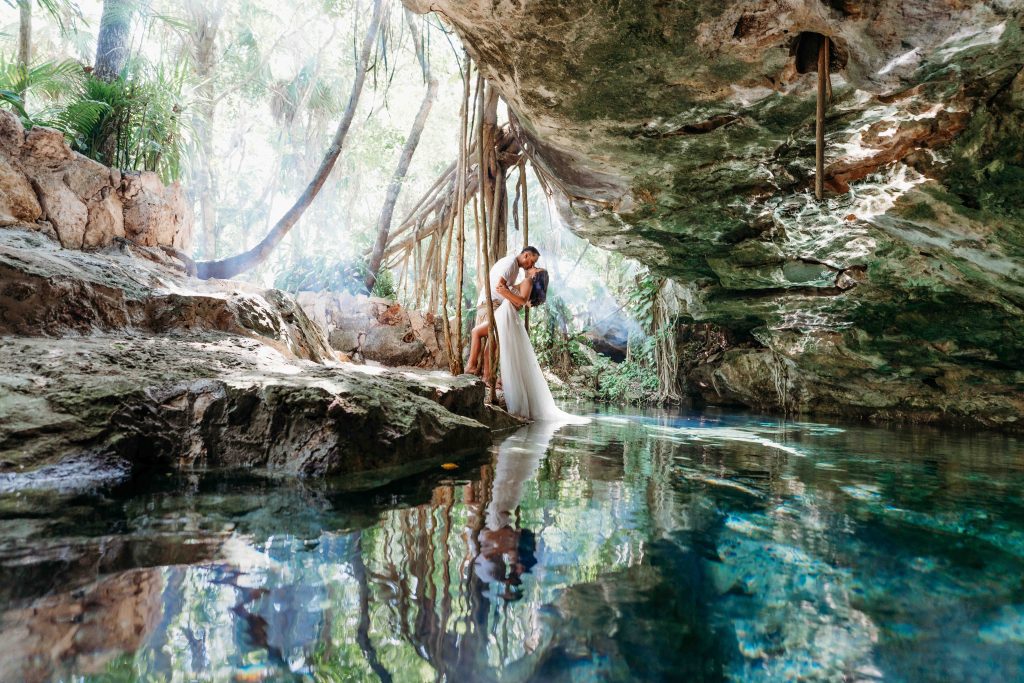 Bride and groom embrace under a rocky overhang beside a clear blue cenote, surrounded by lush greenery and sunlight filtering through trees. Their reflection is visible in the still water. A dream location for wedding photographer cancun