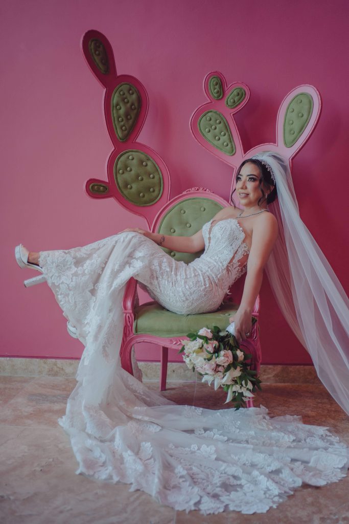 A bride in a white lace gown and long veil sits on a whimsical green and pink chair shaped like a cactus, holding a bouquet, against a pink wall.