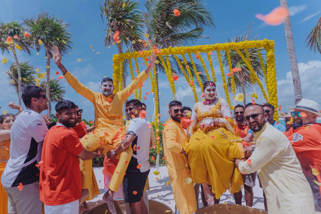 A joyful outdoor celebration with people lifting a man and woman dressed in yellow outfits. Flower petals are thrown in the air, palm trees sway in the background, and a bright yellow floral arch stands behind them.