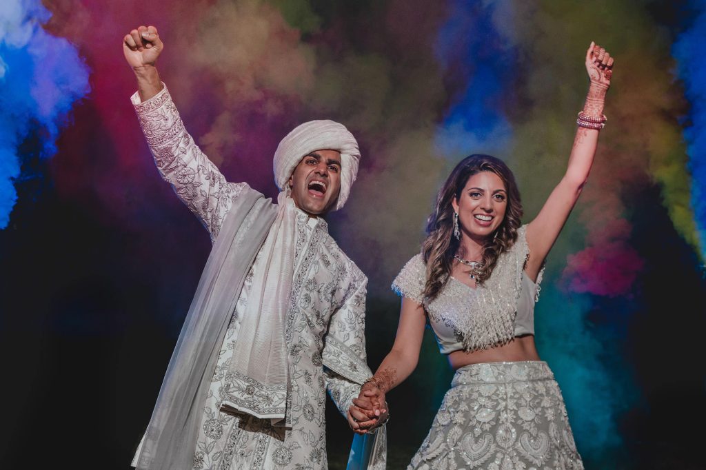 A joyful couple in elaborate white traditional Indian attire hold hands and raise their fists in celebration, captured by a Hotel Xcaret Mexico Beach Wedding Photographer against a colorful, smoky background.